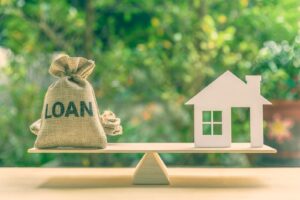 A wooden home and a bag of money with "loan" written on it on a scale